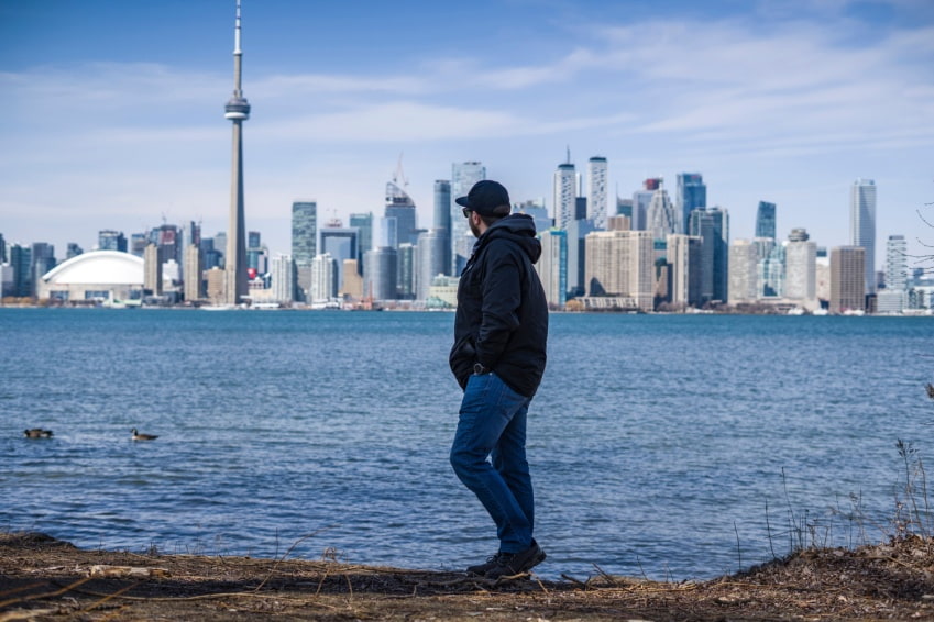 Man posing on the coast of Toronto Island Park with the Toronto skyline, including the CN Tower, in the background across the water.